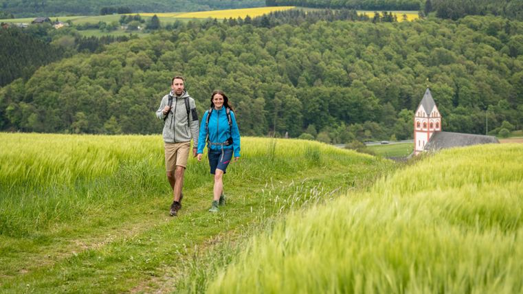 Zwei Wanderer auf einem Pfad durch grüne Felder mit einer Kirche im Hintergrund.
