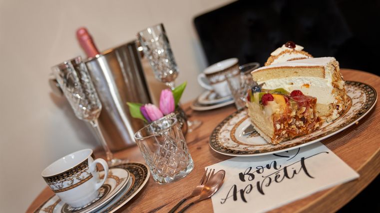 A table setting with a delicious cake, elegant dishes, and champagne glasses. A small flower decoration and a napkin with the inscription "Bon Appétit" complete the picture.