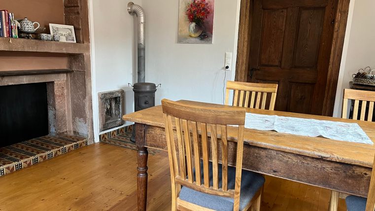 Ein gemütlicher Wohnraum mit einem Holz Tisch und Stühlen. Im Hintergrund sieht man einen Kamin und eine Holztür.