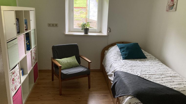 Een eenvoudige slaapkamer met een bed, een stoel en een plank. Het raam laat veel daglicht binnen en er staat een kleine plant op de vensterbank.