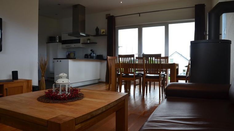 A modern living kitchen with a dining table and many windows. The space is bright and cozy with wood and brown tones.