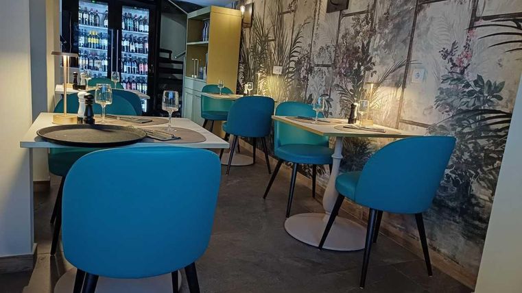A modern restaurant with blue chairs and elegant tables. The walls are decorated with plant motifs and a shelf with drinks is visible.