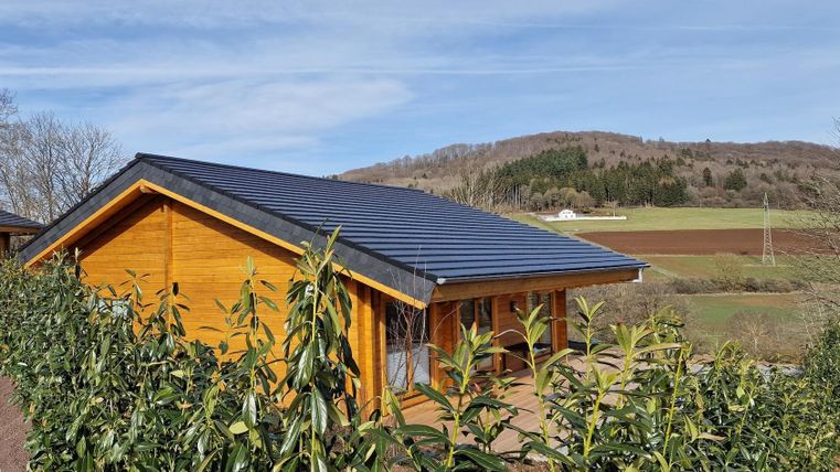 Ein Holzhaus umgeben von grünen Hecken und mit einem schönen Ausblick auf die hügelige Landschaft. Der Himmel ist klar und blau.