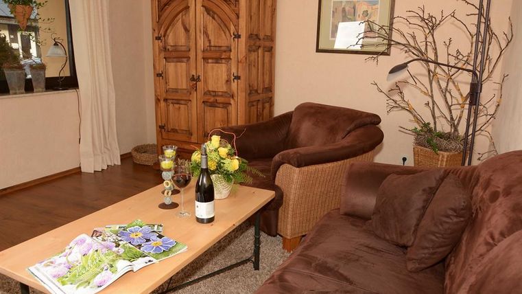 A cozy living room with a dark couch and a wicker chair. On the table, there’s a bottle of wine and fresh flowers.