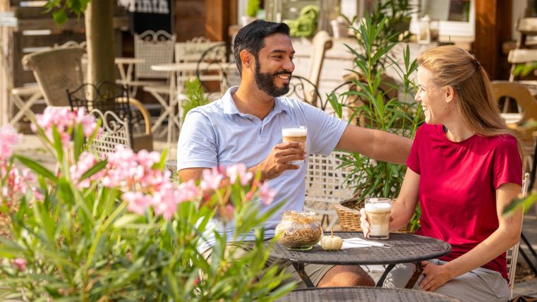 A smiling couple is sitting at an outdoor table, enjoying drinks. Surrounding them are blooming plants, and there is a relaxed atmosphere.