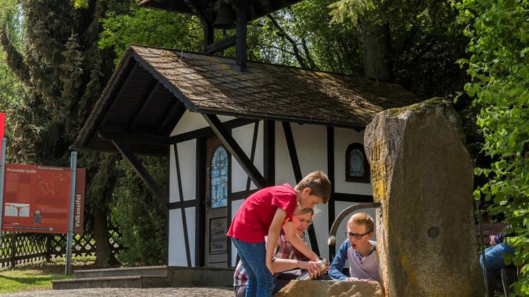 Kinder spielen an einem Stein vor einem kleinen, traditionellen Häuschen im Freien. Die Umgebung ist grün und einladend.