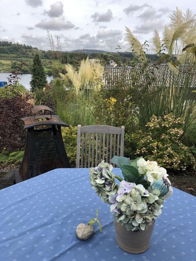 Een mooie tafel met een blauw tafelkleed en een bloempot staat in een tuin. Op de achtergrond zijn groene planten en een meer te zien.