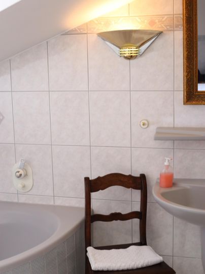 Ein Badezimmer mit einer Badewanne, einem Waschbecken und einem Holzstuhl. An der Wand hängt ein Spiegel und eine Lampe sorgt für Beleuchtung.