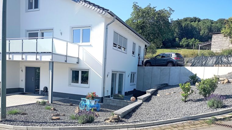 Een modern huis met een verzorgde tuin en grindperceel. Een auto staat in de oprit, en op de achtergrond zijn bomen te zien.