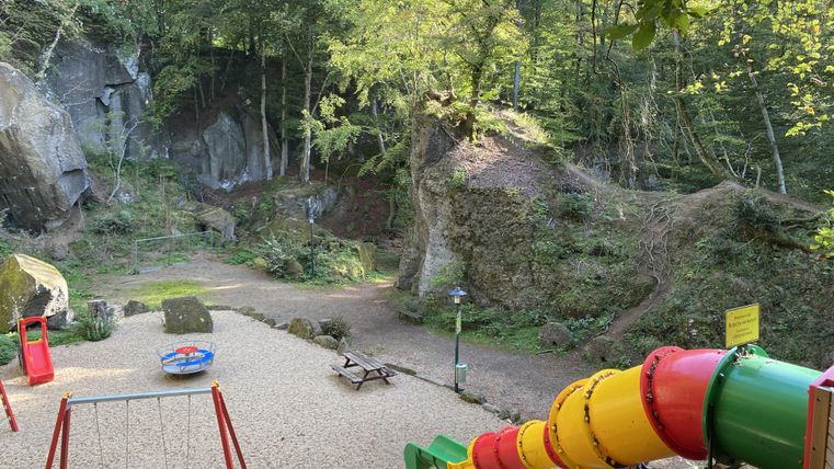 Playground with colorful slide, swing, and climbing frame in a rocky, wooded environment.