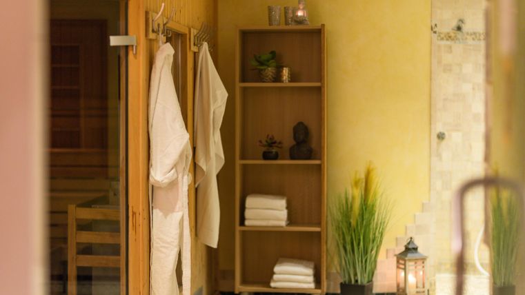 Ein ruhiger Wellnessbereich mit einem Holzregal, sauberen Handtüchern und einem gemütlichen Ambiente. An der Wand hängen Bademäntel und eine Pflanze sorgt für eine entspannende Atmosphäre.