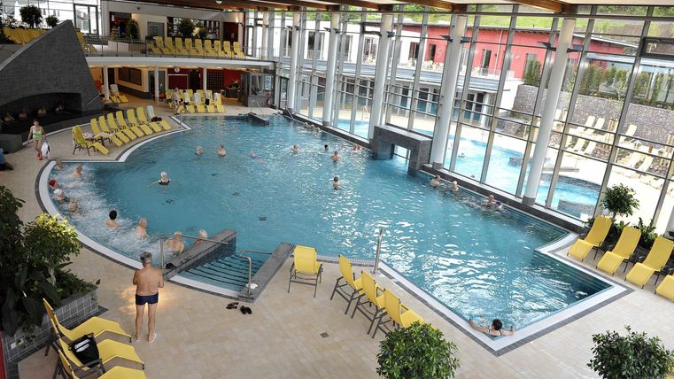 Een inmiddels modern binnenzwembad met blauw water en gele ligstoelen. Door grote ramen stroomt er veel daglicht de ruimte binnen.