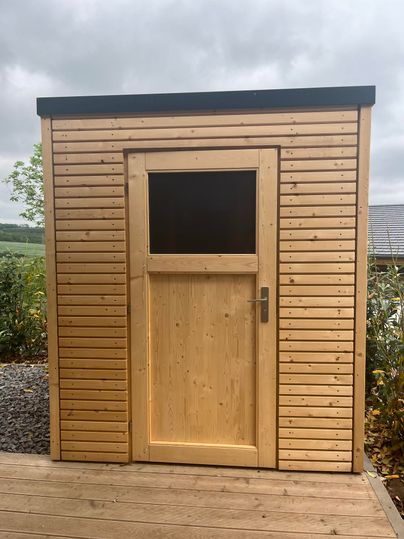 Ein kleines Holzhaus mit vertikalen Holzverkleidungen und einer Fensteröffnung. Es steht in einer grünen Umgebung mit Bäumen im Hintergrund.