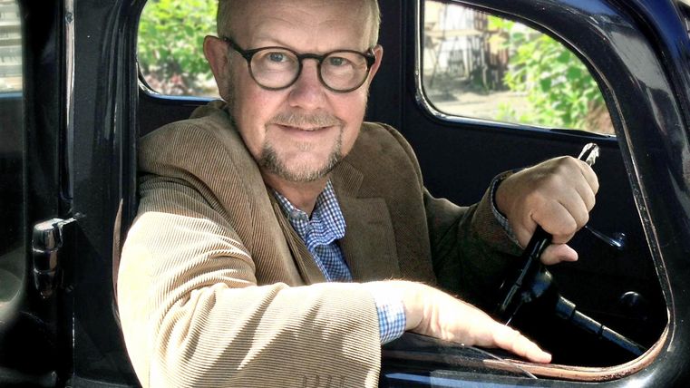 Person mit Brille und Bart sitzt in einem Oldtimer, trägt ein braunes Sakko und lächelt in die Kamera.