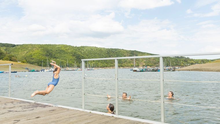 Ein Kind springt von einem Steg ins Wasser, während andere Personen schwimmen. Im Hintergrund sind grüne Hügel und ein klarer Himmel zu sehen.