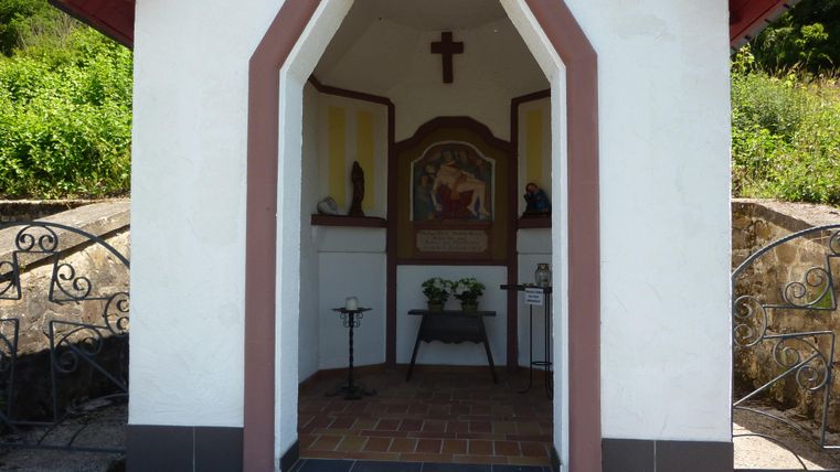 Eine kleine Kapelle mit einem Kreuz und einem Fresko im Inneren. Vor der Kapelle sind Blumen auf einem Tisch platziert.