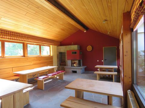 Ein gemütlicher Raum mit Holzdecke und roten Wänden. Zwei große Tische mit Bänken und ein Kamin schaffen eine einladende Atmosphäre.