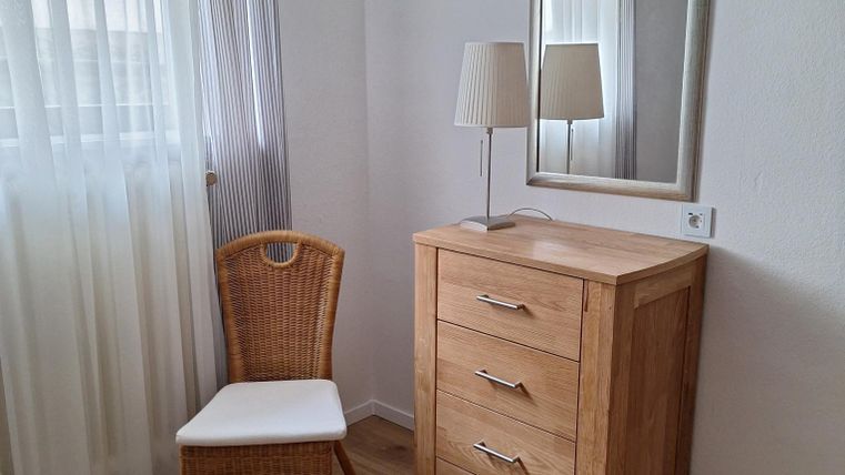 Een lichte kamer met een houten kast en een spiegel. Naast de kast staat een stoel en een lamp.