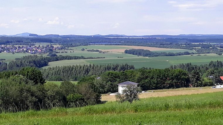 A vast landscape with green fields and hills. In the foreground stands a small building, surrounded by trees and meadows.