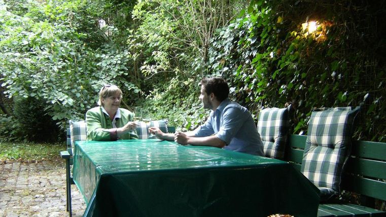 Een gezellige tuin met een tafel en twee personen die met elkaar praten. De omgeving is groen en omgeven door planten.