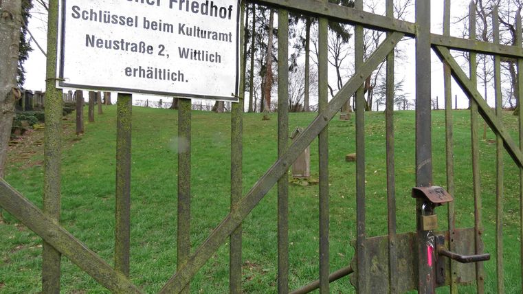 Ein geschlossener Eingang zu einem jüdischen Friedhof mit einem Schild. Die Umgebung besteht aus Rasen und Bäumen im Hintergrund.