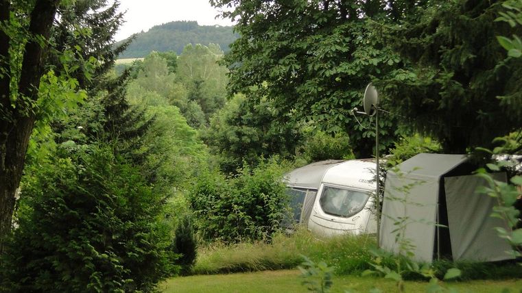 Ein ruhiger Campingplatz umgeben von grüner Natur. Im Hintergrund sind Bäume und leicht bewaldete Hügel zu sehen.