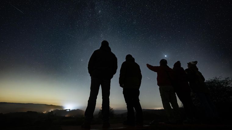 A group of people stands under a clear night sky, gazing at the stars. In the background, a soft light sky is visible.