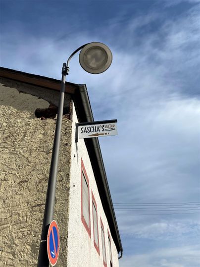 A street sign with the inscription "Sachsenstraße" is attached to a house wall. In the background, a streetlamp and a blue sky can be seen.