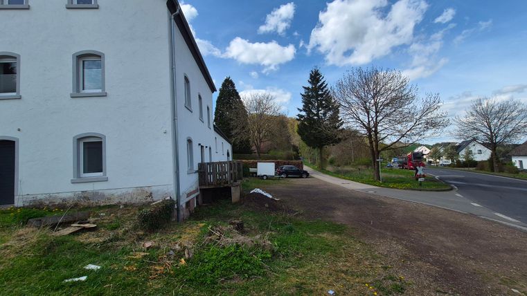 Ein weißes Gebäude an einer Straße, umgeben von Bäumen und einem grünen Bereich. Der Himmel ist klar mit einigen Wolken.