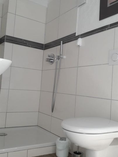 Ein modernes Badezimmer mit einer Dusche und einer Toilette. Die Wände sind hell gefliest und es gibt einen Holzfußboden.