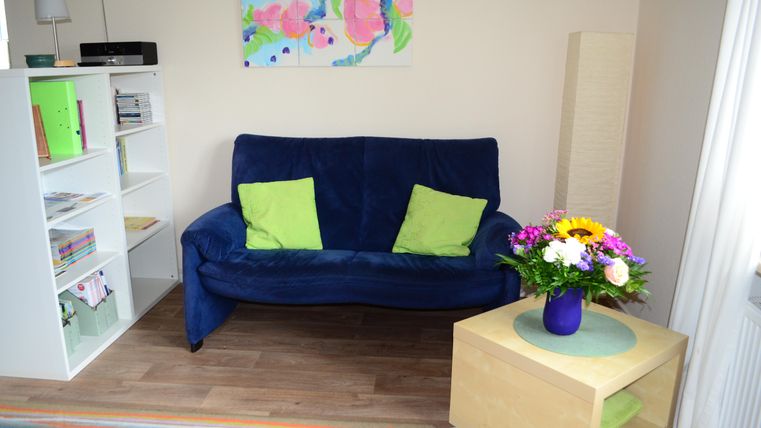 A cozy living space with a blue sofa and light green cushions. The table has a flower vase and in the background there is a bookshelf.