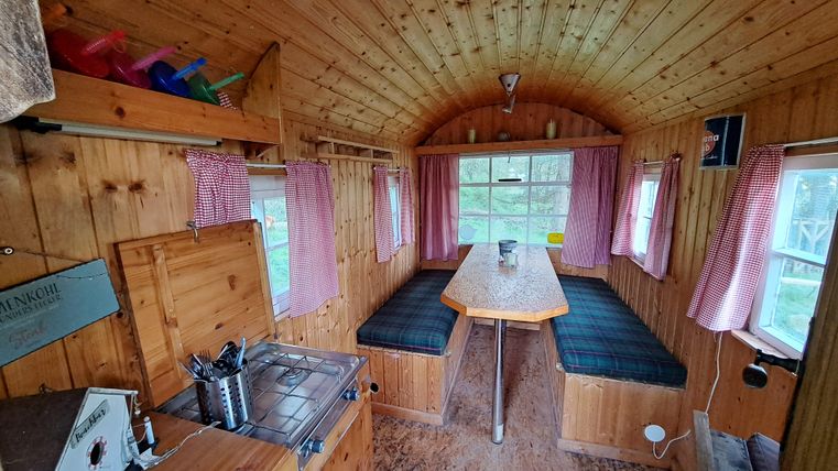 A cozy interior with wooden paneling and a large table in the center. The windows are fitted with curtains and provide plenty of daylight.