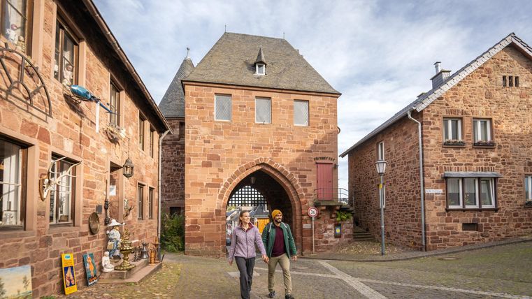 Ein gepflasterter Weg in einer malerischen Stadt mit historischen Gebäuden. Zwei Personen spazieren entspannt durch das Tor.