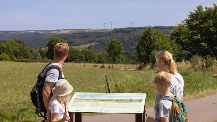 Een gezin staat voor een informatiebord in een groene landschap. Op de achtergrond zijn heuvelachtige velden en bomen te zien onder een heldere lucht.