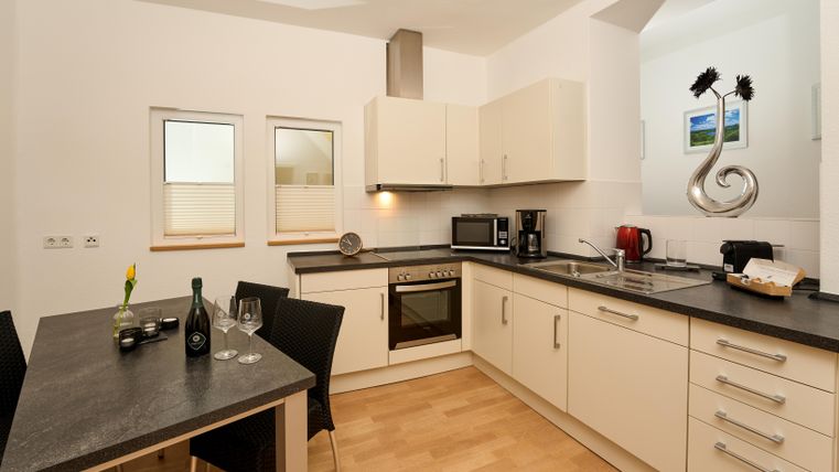 Eine moderne Küche mit hellen Schränken und einer dunklen Arbeitsplatte. Ein Tisch mit Stühlen und einige Küchengeräte sind sichtbar.