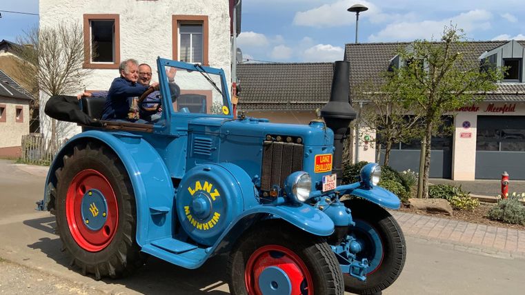 Een blauwe oldtimer tractor staat op een weg. Twee personen zitten vrolijk op de tractor.