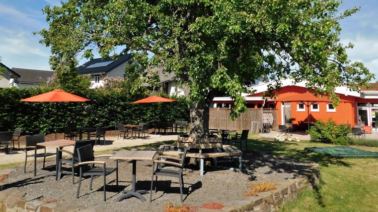 Een uitnodigende buitenruimte met een grote boom en meerdere tafels. Oranje parasols bieden schaduw voor de gasten.