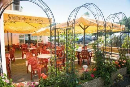 Ein gemütlicher Außenbereich eines Restaurants mit roten Tischen und Stühlen. Gelbe Sonnenschirme spenden Schatten und Blumen schmücken die Umgebung.