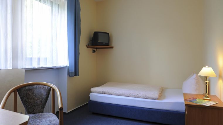 Ein einfaches Zimmer mit einem Einzelbett, einem Tisch und einem Stuhl. Es gibt ein Fenster mit viel Tageslicht und einen Fernseher.