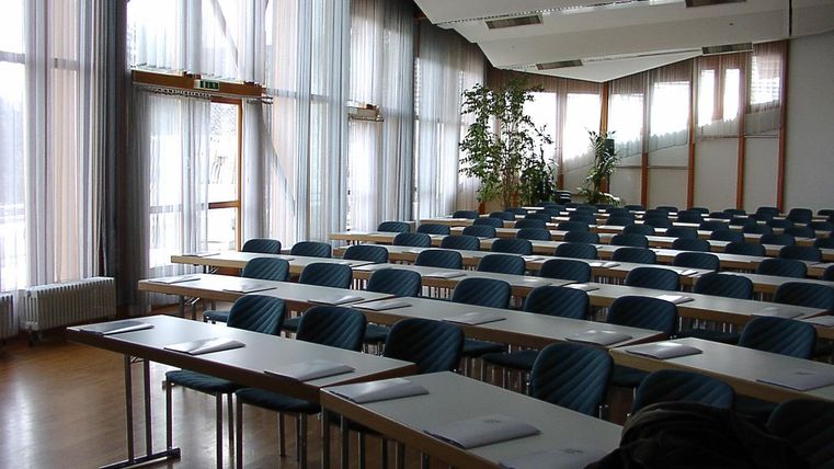 Ein großer, leerer Seminarraum mit Reihen von blauen Stühlen und Tischen. Viel Tageslicht strömt durch die großen Fenster.