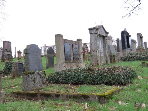 Ein alter Friedhof mit verschiedenen Grabsteinen und viel grünem Gras. Es ist bewölkt und sieht unheimlich still aus.
