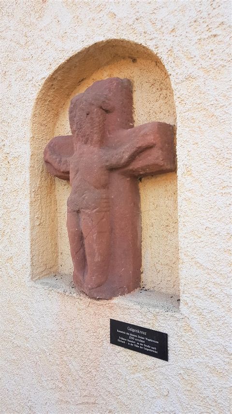 Eine terrakottafarbene Statue eines Kreuzes ist in einer Wandnische angebracht. Darunter befindet sich ein kleiner Schild mit weiteren Informationen.