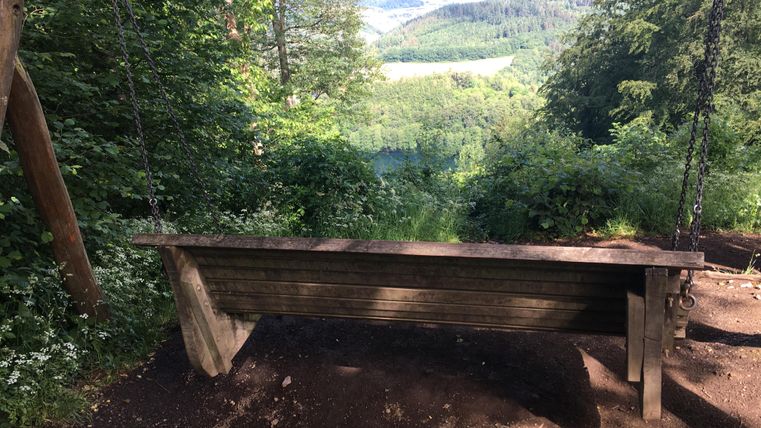 A wooden bench stands at a viewpoint surrounded by trees. In the background, a beautiful landscape with hills and a clear sky can be seen.