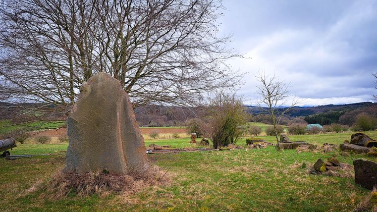 Een grote stenen hoop op een groene weide onder een bewolkte lucht. Op de achtergrond zijn verspreide bomen en heuvels te zien.