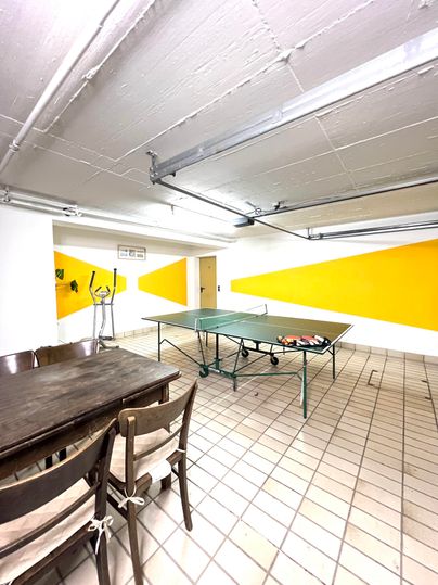 A bright room with a table for table tennis and a large wooden table. The walls are partially painted yellow and there are a few chairs.