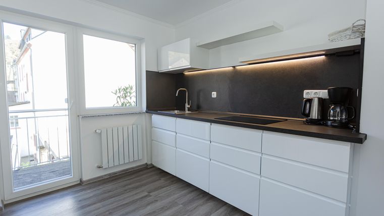 Eine moderne Küche mit weißen Schränken und einem dunklen Arbeitsbereich. Große Fenster sorgen für viel Tageslicht.