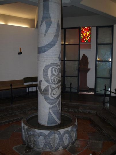 Ein dekorierter Säulenbrunnen in einem Innenraum. Farbiges Glasfenster und eine Statue sind im Hintergrund sichtbar.