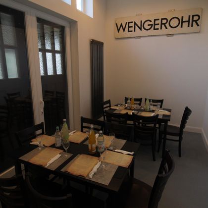 A restaurant with dark wooden tables, set with glasses and drinks. A sign reading 'WENGEROHR' hangs on the wall.