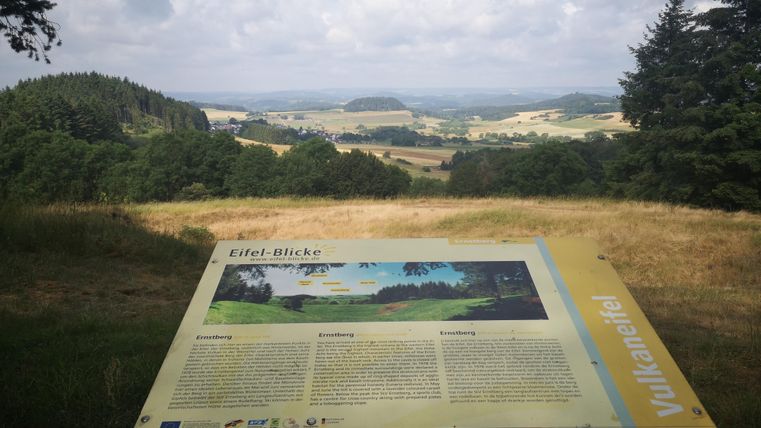 Een uitzichtpunt met informatie over de Eifel. Op de achtergrond strekken zich groene heuvels en een bewolkte lucht uit.
