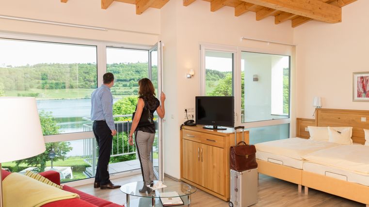 Ein modernes Hotelzimmer mit großen Fenstern und Blick auf die grüne Landschaft. Zwei Personen stehen am Fenster und genießen die Aussicht.